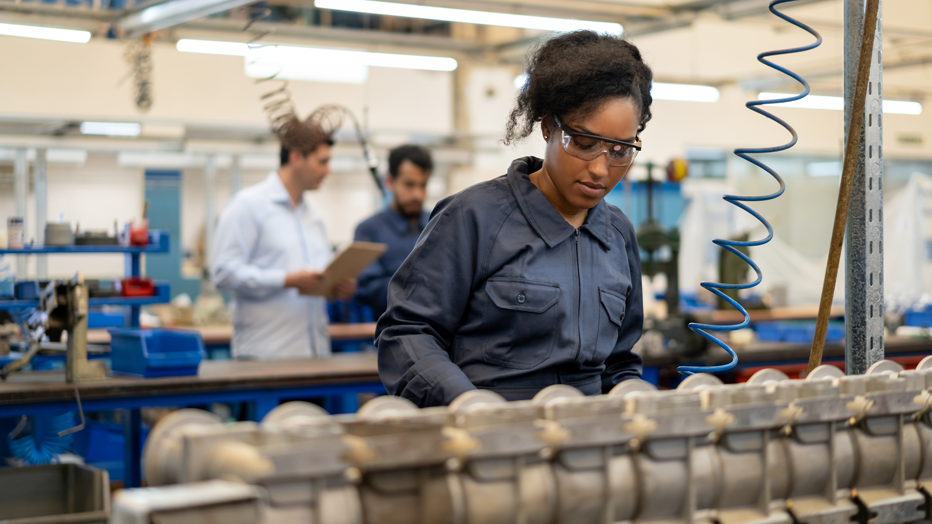 Une travailleuse en combinaison s'affaire avec application à son poste sur une chaîne de fabrication, plus loin, un manager discute avec un autre employé.