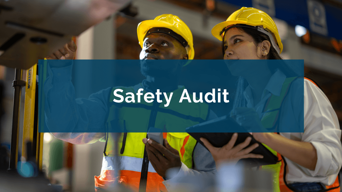 Two auditors wearing hard hats and safety vests, inspecting a machine, one holding a pencil and a clipboard