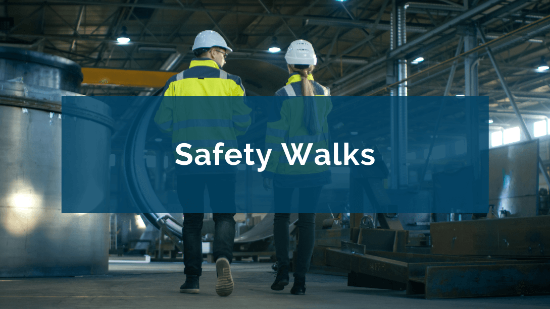 Two workers wearing hard hats and safety vests, seen from behind, walking across a factory floor